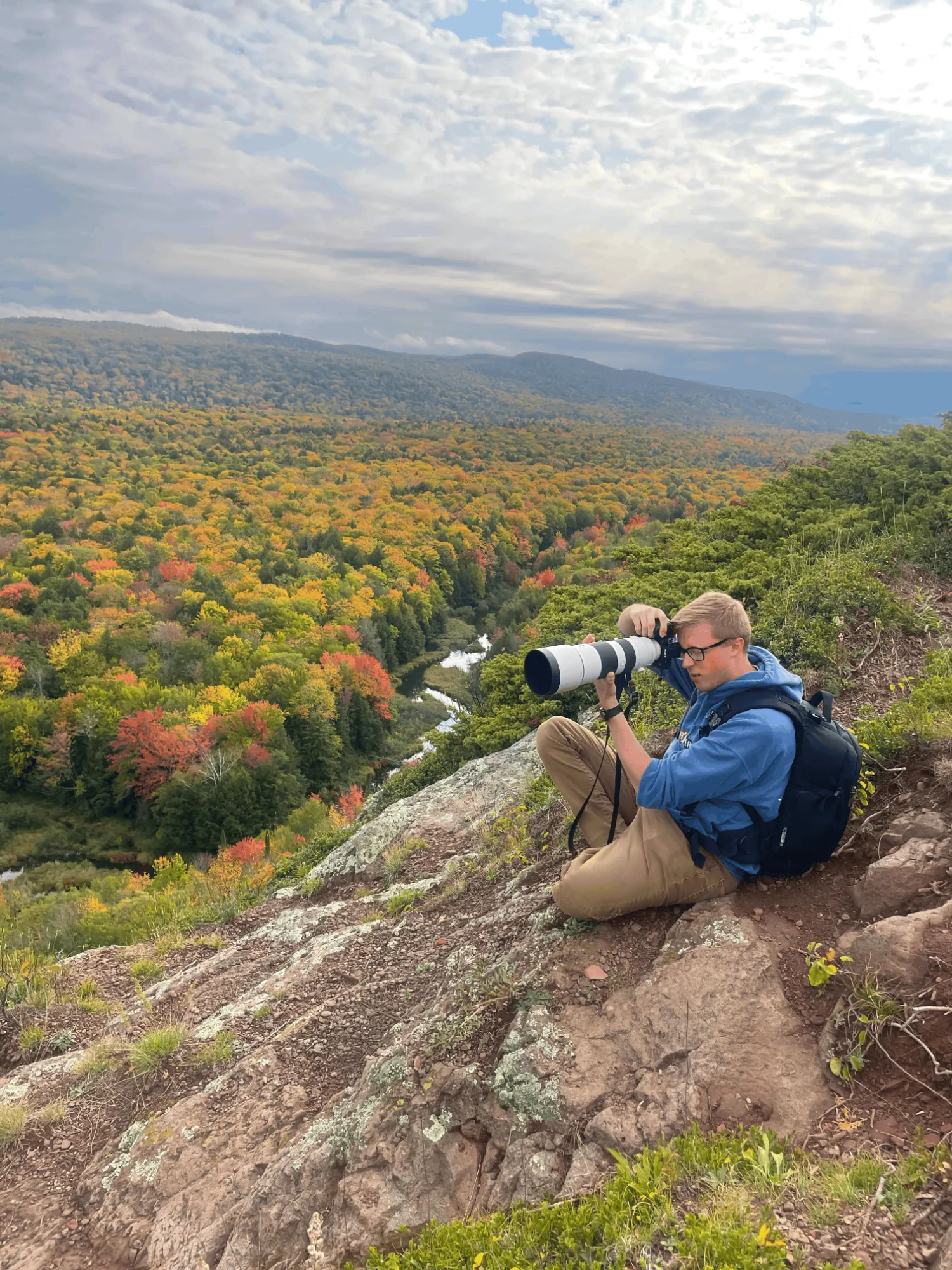Lucas sitting on a rocky overlook in autumn, shooting the forested valley below with a long telephoto lens.